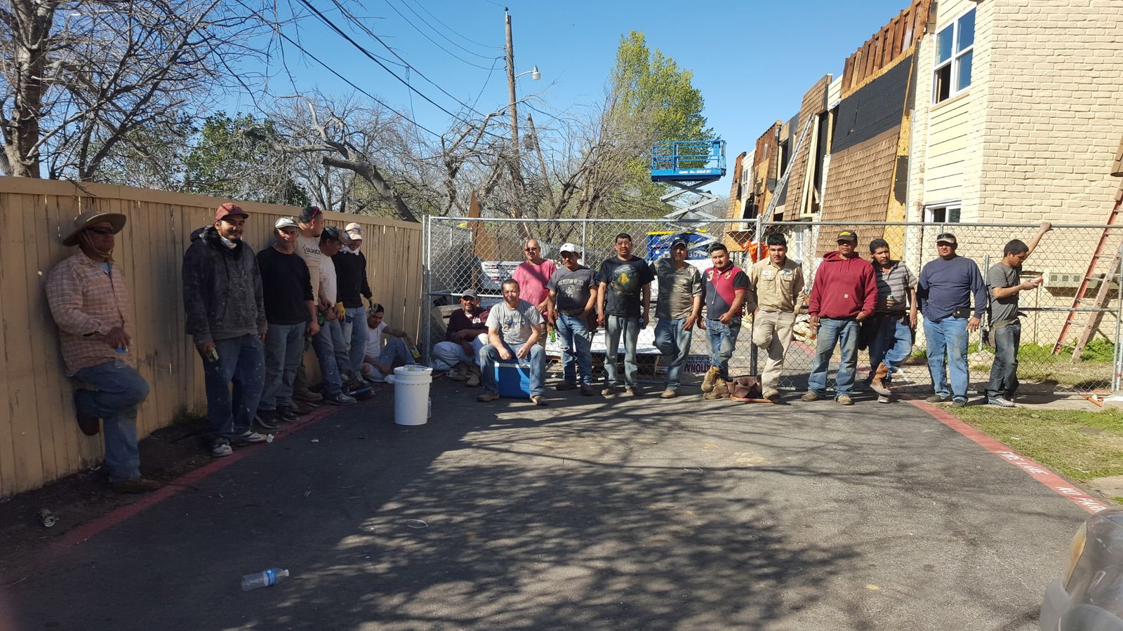 More than twenty workers lined up in a driveway beside a fence at an apartment complex under renovation