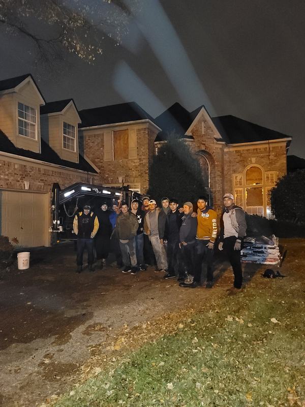 Crew standing together after dark in front of a brick home with work lights aimed at the roof and a trailer in the driveway
