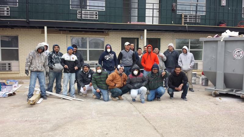 Group of workers in hooded sweatshirts and dust masks posing in front of a two-story building and a roll-off dumpster