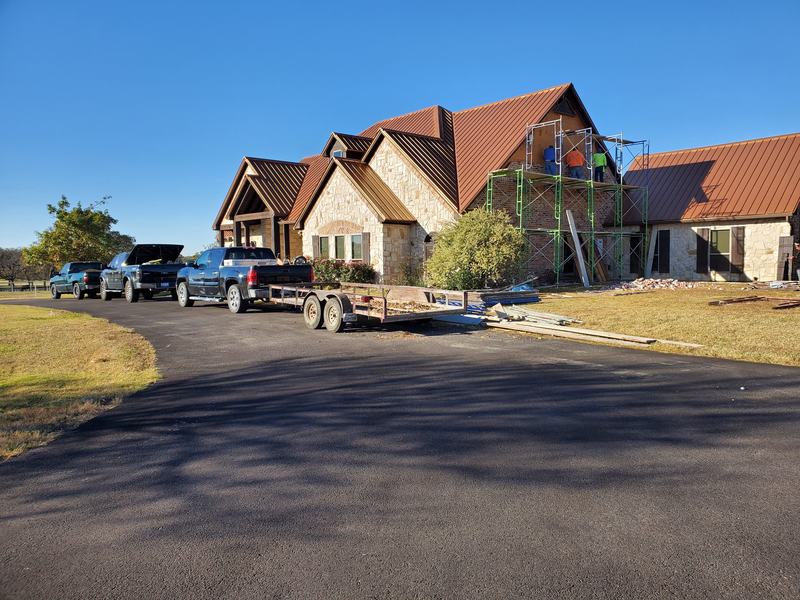 Stone and brick home with a rust-red metal roof, scaffolding at the front gable and work trucks and a trailer in the driveway