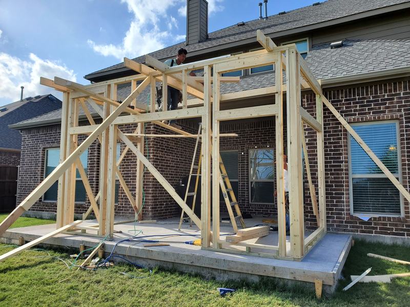 Wood framing for a backyard addition on a concrete slab with workers on the structure and lumber on the lawn